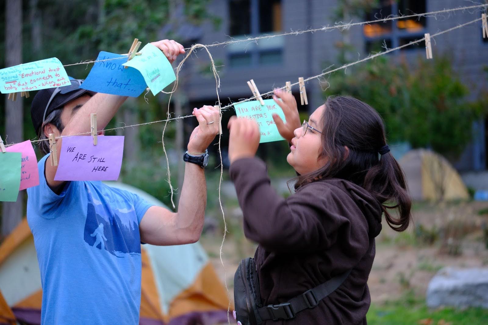 Two people stand at and actively engage with what appears to be a clothesline. Colourful pieces of paper
with text written on them are pinned to the clothesline. On one piece of paper can be seen the hand-written
text "WE ARE STILL HERE" while on others can be seen the words "Know that..." and "You are your ancestors".