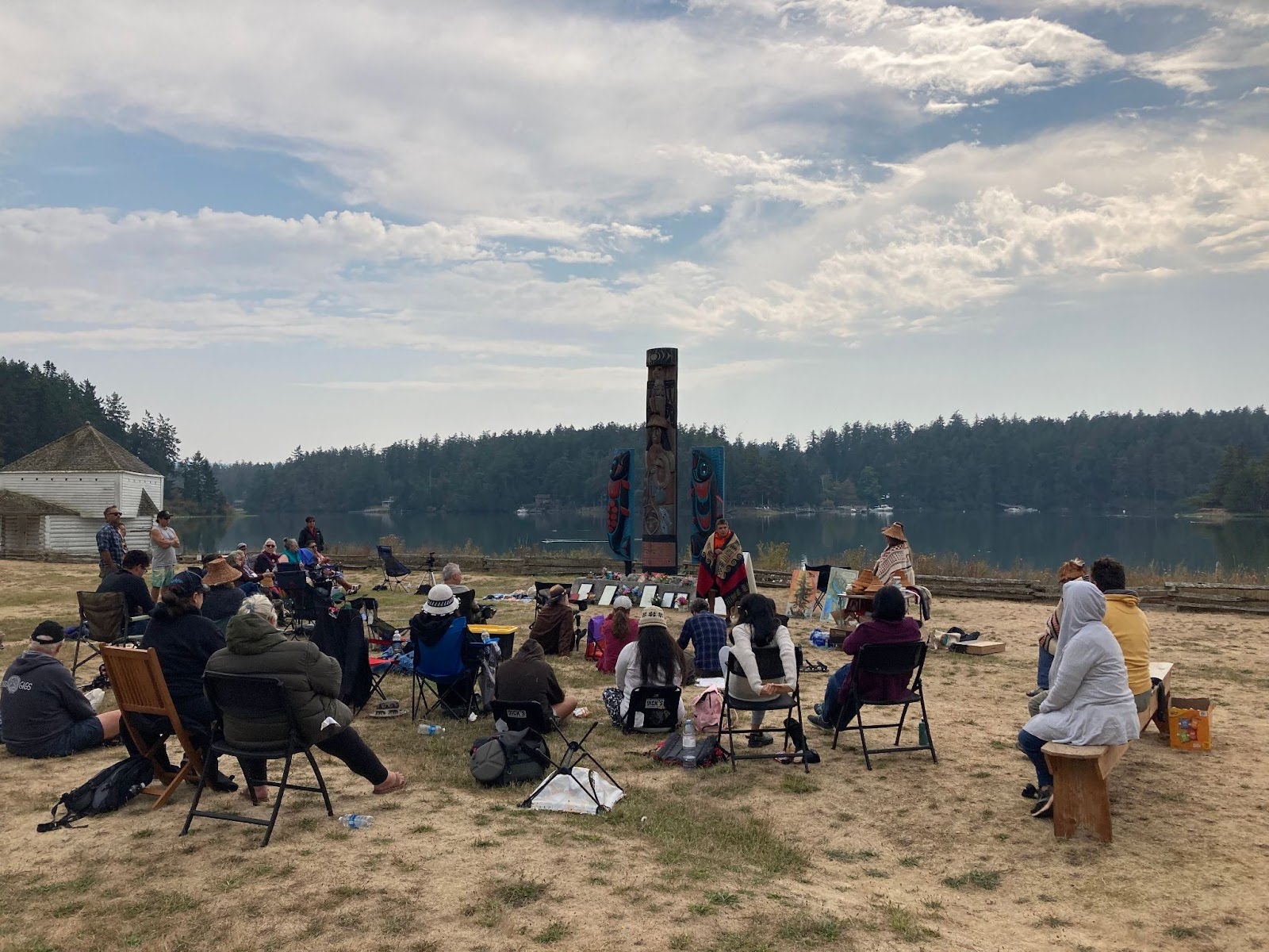 A group of approximately 25 people stand and sit in a grassy area by the water in front of 3 carved and
painted Strait Salish story poles. A man wearing a blanket over his shoulders stands in front of the group and
appears to be talking, while a woman in a woven Salish blanket and cedar hat stands near him listening.
A white clapboard building can be seen in the background.