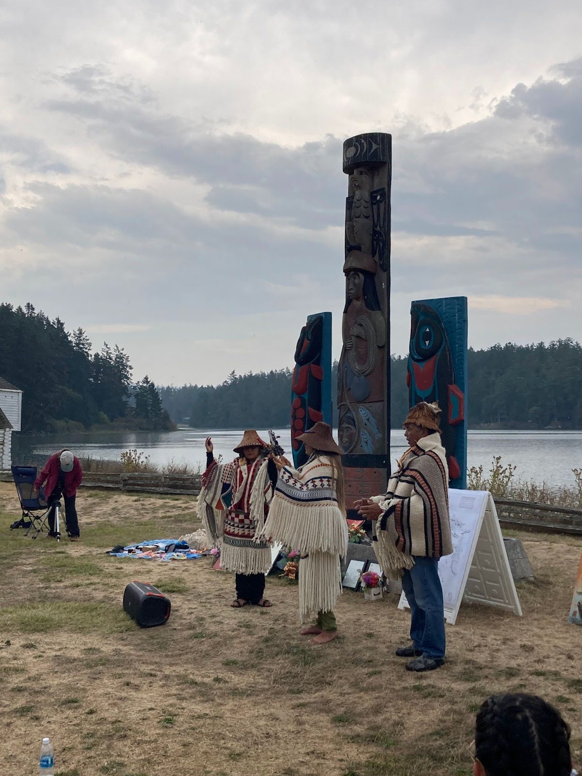Three people wearing Coast Salish woven clothing and cedar hats stand in front of 3 story poles with their
hands raised in front of their bodies. One of them holds a rattle in each hand.
In the background water and trees can be seen.